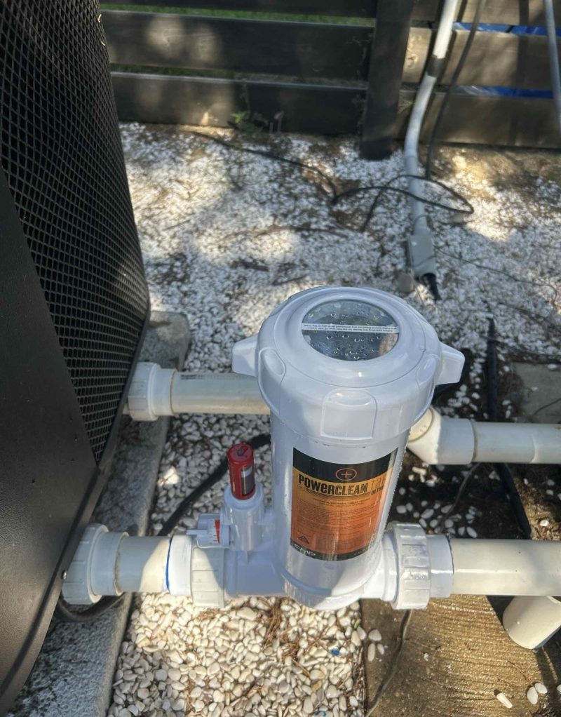 White pool filtration pump with PowerClean label, connected to PVC pipes outdoors on gravel.